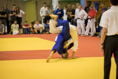 Zwei Judoka auf gelber Matte in einer Wurfposition; Zuschauer und ein Kampfrichter stehen drumherum.