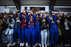 Menschenmenge mit erhobenen Händen in einer überdachten Tribüne; einige tragen blaue Overalls und Schals.