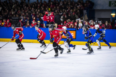 Eishockeyspiel in der Halle: mehrere Spielerinnen in roten und blauen Trikots auf dem Eis, Zuschauer im Hintergrund.