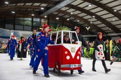 Kostümierte Personen ziehen auf einer Eisfläche einen rot-weißen Wagen bei einer Karnevalsveranstaltung.
