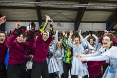 Gruppe von Menschen in bunten Kostümen tanzt und feiert in einer Halle vor großem Publikum.