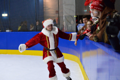 Person im Weihnachtsmannkostüm auf einer Eisbahn neben der Bande, rechts Zuschauer hinter der Absperrung.