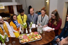 Sieben Personen sitzen um einen runden Tisch mit belegten Brötchen, Flaschen, Gläsern und weißen Blumen.