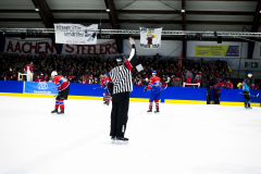 Eishockeyspiel mit Schiedsrichter auf dem Eis, Spielern vor der Bande und Zuschauern auf der Tribüne.