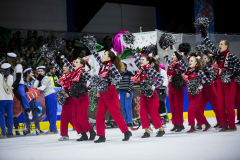 Eisläuferinnen in roten Hosen und karierten Hemden mit Pompons auf dem Eis; dahinter weitere Personen.
