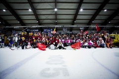 Große Gruppe verkleideter Personen auf einer Eisfläche in einer Halle, einige mit Helmen, Fahnen und Pompons.
