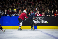 Zwei Eishockeyspieler an der Bande vor Publikum, schwarzer Puck auf dem Eis.