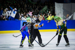 Eishockeyszene mit Schiedsrichter und drei Spielerinnen auf dem Eis, Zuschauer im Hintergrund.