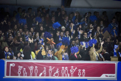 Fans mit blauen Pompons und Schals stehen und jubeln hinter einer roten Absperrung.