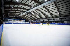 Eishalle mit Eisfläche, mehreren Spielern, Zuschauern auf der Tribüne und hoher Hallendecke.