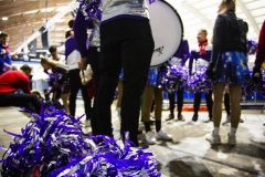 Person mit Trommel und violetten Pompons in einer Halle, weitere Personen im Hintergrund.