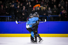 Zwei Eishockeyspieler in blauen und schwarzen Trikots umarmen sich auf dem Eis, Publikum im Hintergrund.