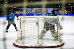 Eishockeytor mit Torwart im blauen Trikot und Spieler links auf dem Eis in einer Halle.