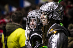 Person in silberner Schutzkleidung und Helm mit Gittermaske, Vorderansicht, unscharfe Personen im Hintergrund.