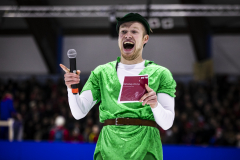 Person in grünem Kostüm hält ein Mikrofon und ein Heft mit der Aufschrift „Eishockey Uni-Cup“.