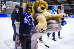 Menschen auf einer Eisfläche tragen ein großes Teddybär-Kostüm; eine Person hält ein hohes braun-gelbes Schild hoch.