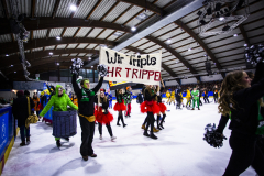 Personen auf einer Eisbahn, eine hält ein Schild mit „Wir Triple HR TRIPPEL“, andere mit Pompons und Kostümen.