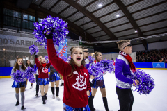 Mehrere Personen mit lila Pompons auf einer Eisfläche in einer Halle.