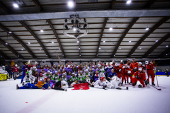 Gruppenfoto von vielen Menschen in bunten Kostümen auf einer Eisfläche in einer Halle, mit Beleuchtung an der Decke.