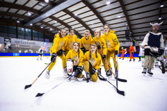 Sechs Personen in gelben Hockey-Outfits posieren auf einer Eishockeyfläche, halten Schläger; weitere Spieler im Hintergrund.