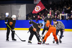 Eishockeyspiel mit vier Spielern und einem Schiedsrichter auf dem Eis; im Hintergrund sitzt Publikum und eine rote Fahne.
