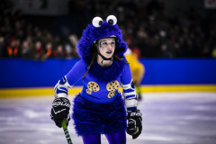 Person im blauen Monsterkostüm mit Eishockeyausrüstung auf dem Eis, Zuschauer im Hintergrund.