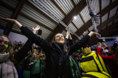 Menschenmenge in einer Halle, eine Person mit ausgebreiteten Armen im Vordergrund, rechts eine Person in gelb-schwarzem Kostüm.