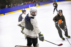 Mehrere Eishockeyspieler auf einer Eisfläche; einer im Vordergrund mit weißem Helm und dunklen Flügeln auf dem Rücken.