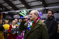 Mann mit Mikrofon vor Gruppe mit Pompons und Flagge in einer Halle