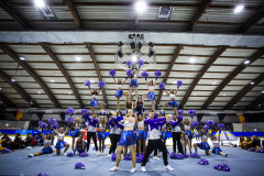 Cheerleadergruppe in blauen Uniformen mit violetten Pompons in einer Turnhalle, in Pyramiden posierend.