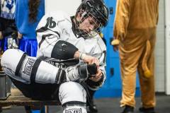 Eishockeyspieler im Umkleidebereich beim Schnüren der Schlittschuhe, während im Hintergrund verkleidete Personen stehen.