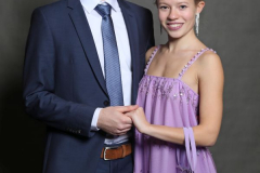 Mann im blauen Anzug und Frau im lilafarbenen Kleid stehen vor grauem Hintergrund und halten sich an den Händen.