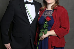 Zwei Personen in Abendkleidung vor grauem Hintergrund; eine Person hält eine rote Rose.