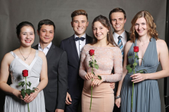 Sechs festlich gekleidete Personen stehen nebeneinander und halten rote Rosen.