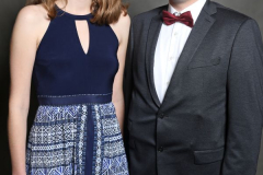 Zwei Personen stehen nebeneinander vor grauem Hintergrund; links im blauen Kleid, rechts im dunklen Anzug mit Fliege.