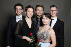 Fünf Personen in festlicher Kleidung vor dunklem Hintergrund, zwei halten rote Rosen.