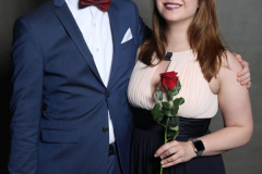 Mann im blauen Anzug und Frau im dunklen Kleid mit roter Rose, stehend vor grauem Hintergrund.