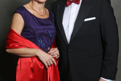 Ein Mann im schwarzen Anzug und eine Frau im violetten Kleid mit roter Stola stehen vor grauem Hintergrund.