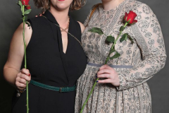 Zwei Frauen stehen nebeneinander vor grauem Hintergrund und halten je eine rote Rose in der Hand.