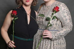 Zwei Frauen stehen nebeneinander vor grauem Hintergrund und halten rote Rosen.