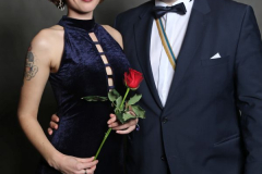 Zwei Personen in festlicher Kleidung, eine hält eine rote Rose, vor grauem Hintergrund.