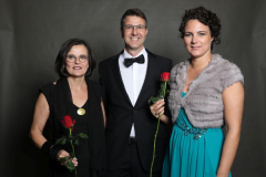 Drei Personen vor dunklem Hintergrund, zwei Frauen und ein Mann in Abendkleidung, jede mit einer roten Rose.