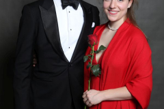 Mann im schwarzen Smoking und Frau im roten Kleid mit Rose, vor dunklem Hintergrund.
