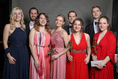 Sechs Menschen in festlicher Kleidung stehen vor grauem Hintergrund; vier halten rote Rosen.