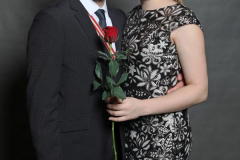 Mann im Anzug und Frau im schwarzen Kleid stehen eng zusammen und halten eine rote Rose.