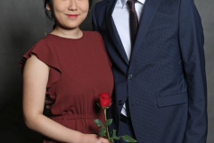 Frau im roten Kleid hält eine rote Rose, neben ihr steht ein Mann im dunkelblauen Anzug vor grauem Hintergrund.