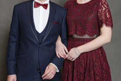 Ein Mann im dunkelblauen Anzug und eine Frau im bordeauxroten Spitzenkleid stehen vor grauem Hintergrund.