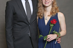 Mann im schwarzen Anzug und Frau im blauen Kleid mit roter Rose vor grauem Hintergrund.