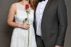 Frau in weißem Spitzenkleid mit roter Rose, neben Mann im dunklen Anzug vor grauem Hintergrund.