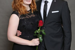 Zwei Personen in schwarzer Kleidung vor grauem Hintergrund; die Frau hält eine rote Rose.